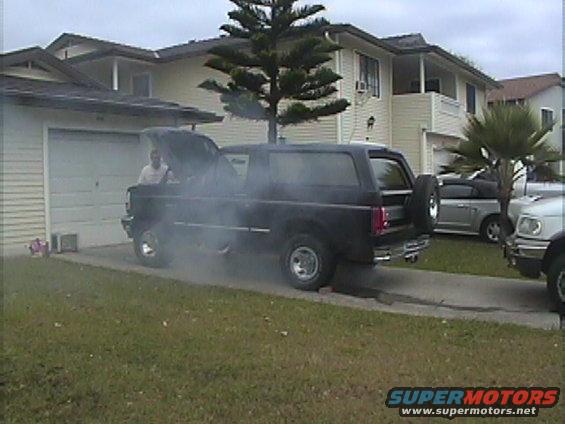 Black SUV with hood open, smoke billowing out, parked in a driveway next to other vehicles
