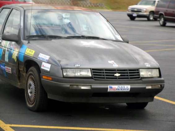 Dark gray sedan with stickers on the hood and side, a Chevrolet emblem on the grille, and a "Peace is Patriotic" bumper sticker
