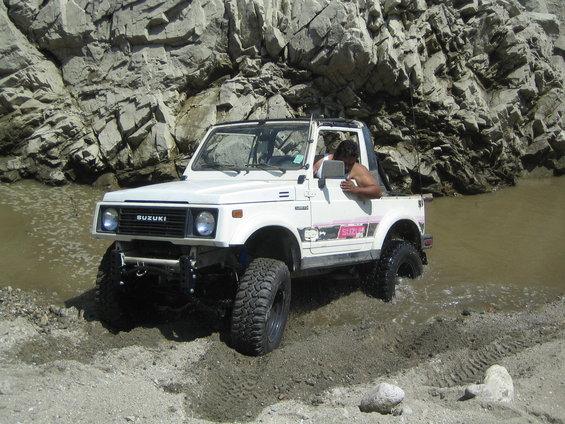 White 1987 Suzuki Samurai driving through shallow water and mud near rocky cliffs