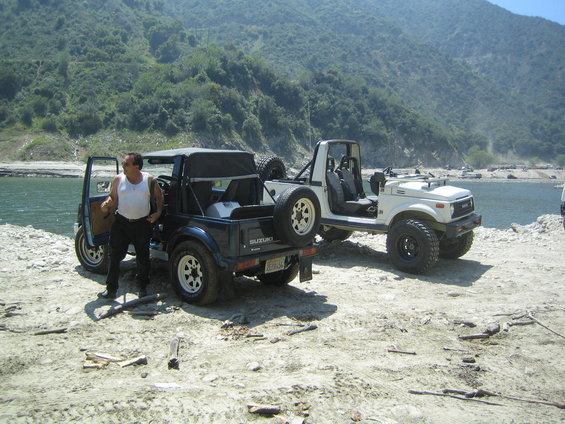 Two 1987 Suzuki Samurai off-road vehicles parked on a sandy shore near a lake