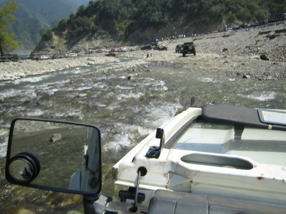 View from inside a white off-road vehicle crossing a shallow, rocky river. Other off-road vehicles are visible on the far bank and in the water