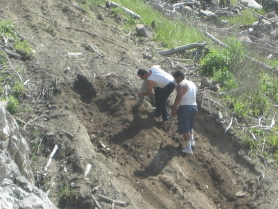 Two men digging on a dirt hillside with scattered debris and vegetation