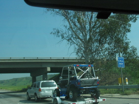 Blue off-road vehicle on trailer being towed by white SUV. Overpass and trees in background