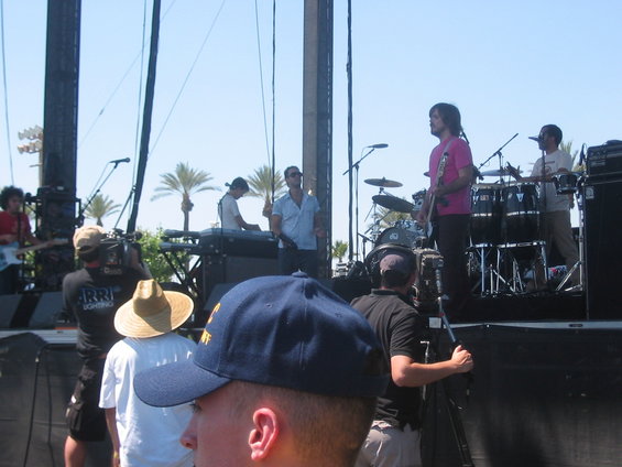 Band playing music on an outdoor stage with palm trees in the background. People are filming the performance