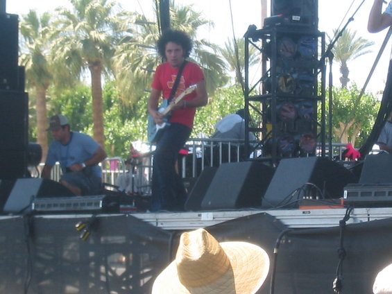 Musicians playing guitars on an outdoor stage with palm trees in the background