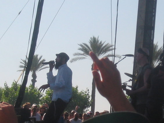 Man with beard and cap singing into microphone on outdoor stage, palm trees behind him