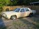 1985 Ford Crown Victoria LTD - A tan sedan with a wooden fence in the background and aftermarket wheels is parked on a gravel surface.