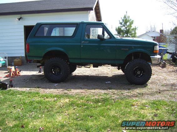 Green 1996 Ford Bronco with a lift kit and large tires, parked on gravel in front of a garage