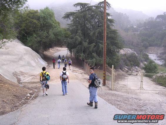 Group of hikers walking on a paved path in a hilly, wooded area with a fence and gate
