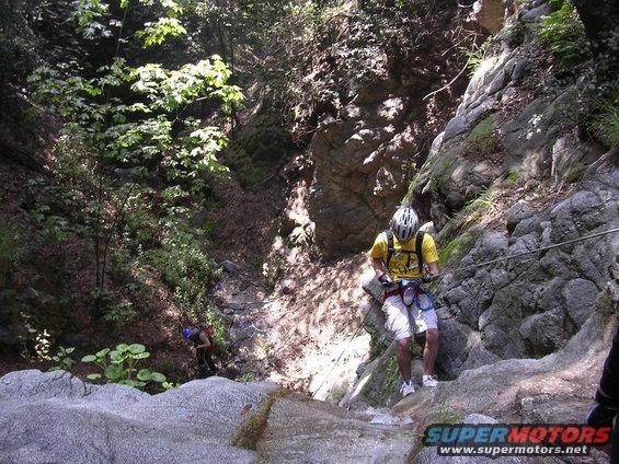 Two people rappelling down a rocky canyon with trees and water
