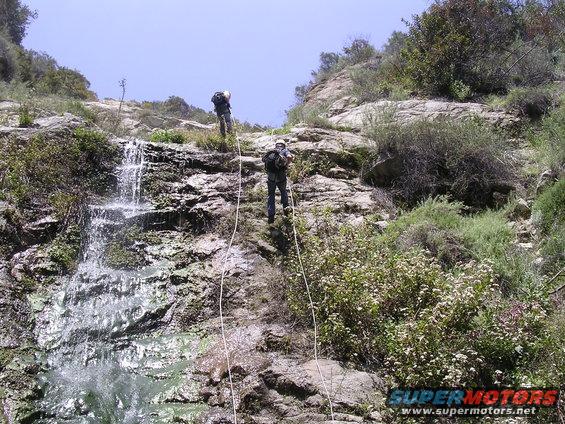 Two people rappelling down a rocky cliff next to a waterfall. They are using ropes and wearing backpacks