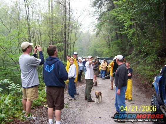 Group of people and off-road vehicles on a dirt road in a wooded area, with a dog on a leash
