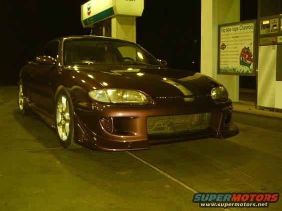 Maroon 1993 Mazda MX-6 coupe with custom body kit and racing stripes parked at a gas station