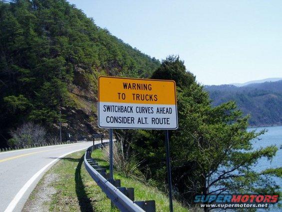 Roadside sign warns trucks of switchback curves ahead and suggests an alternate route. Mountainous terrain and water are visible