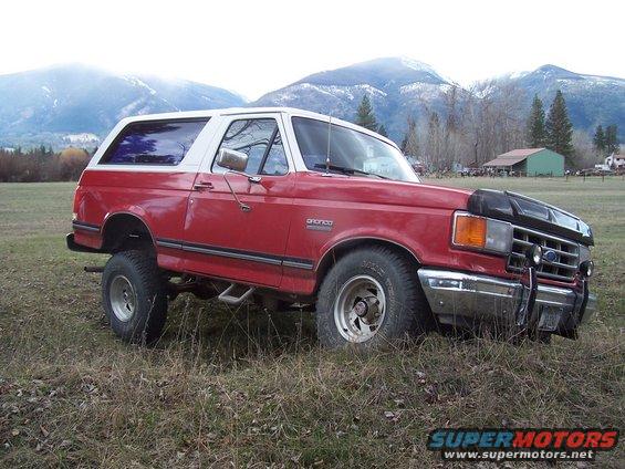 Red 1988 Ford Bronco with white top parked in a grassy field with mountains in the background