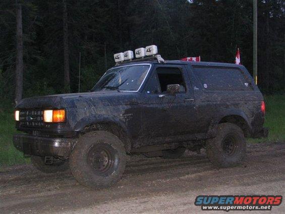 Mud-covered black 1987 Ford Bronco with light bar on roof and Canadian flag