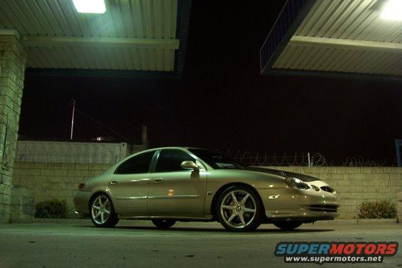 Gold sedan parked at night under a covered area. It has aftermarket wheels and a dark hood