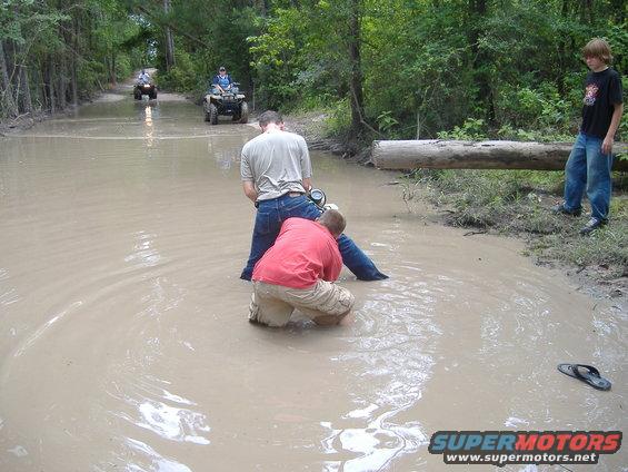 Two people attempt to push an ATV out of a muddy water crossing while two other ATVs are further down the trail
