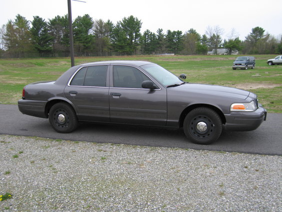 Gray Ford Crown Victoria police car parked on gravel, trees and field in background