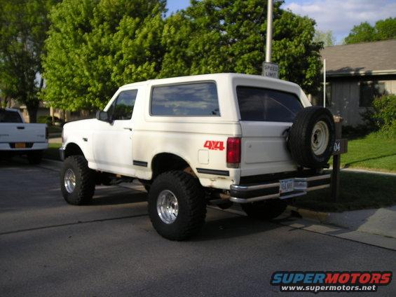 1990 Ford Bronco parked on street, rear view, 4x4 decal visible. White pickup truck in background