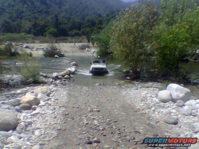 White SUV crossing a rocky riverbed with trees and hills in the background