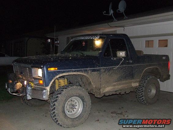 Mud-covered blue Ford Bronco with aftermarket bumper and winch parked in front of a garage