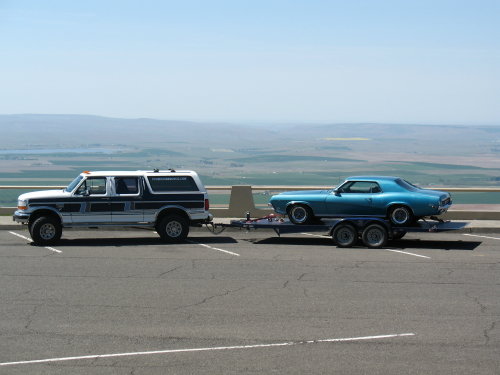 Ford Bronco towing a blue coupe on a trailer, parked on an overlook