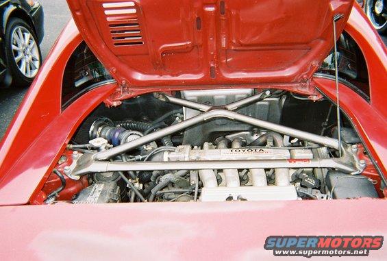 Red Toyota MR2 engine bay with cross brace and Toyota engine cover. Another car is partially visible on the left