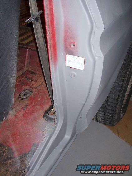 Interior of a vehicle, showing the door jamb area with a seatbelt and a sticker on the door frame. The floor is red and appears rusty