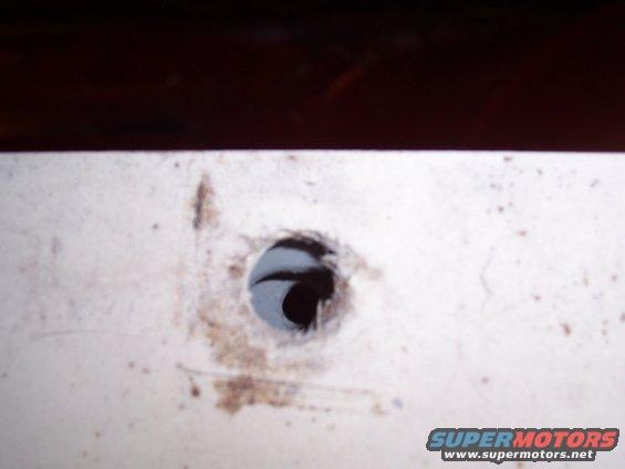 Close-up of a hole drilled into a white panel, with some dirt smudges. Supermotors.net logo visible