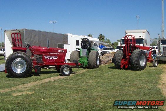 Two modified tractors, one red International and one green, parked on grass with trailers and trucks behind them