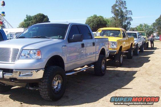 Several lifted pickup trucks parked in a line on a dirt lot, with a Ferris wheel in the background