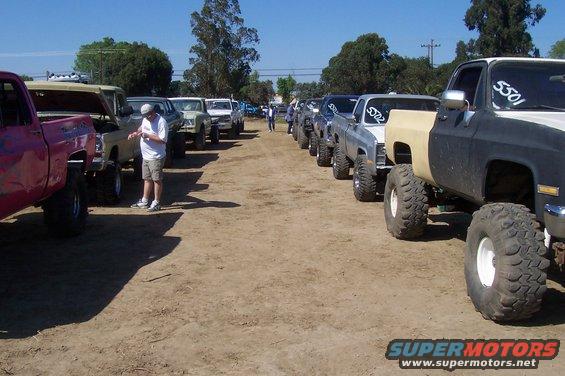 Several lifted pickup trucks parked in a dirt lot, some with hoods open. One truck is black and tan with huge tires