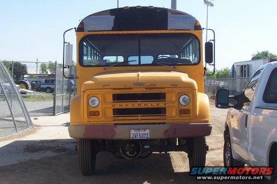 Front view of a yellow Chevrolet bus with a white pickup truck to its right. The bus has a California license plate reading 3VEK899
