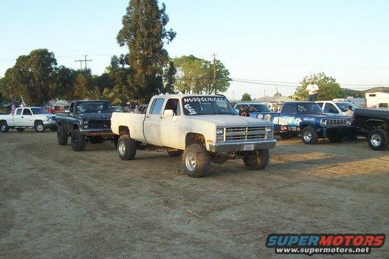 Several lifted pickup trucks parked in a dirt lot, one white crew cab in foreground
