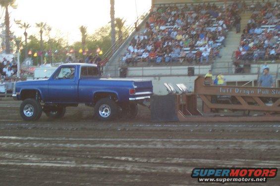 Blue pickup truck with lifted suspension pulling a sled at a tractor pull event. Spectators are in the bleachers