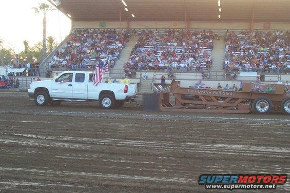 White pickup truck with American flags on the back, towing a large sled on a dirt track in front of a crowd