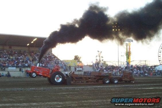 Tractor pulling a sled with black smoke billowing from exhaust, crowd in bleachers