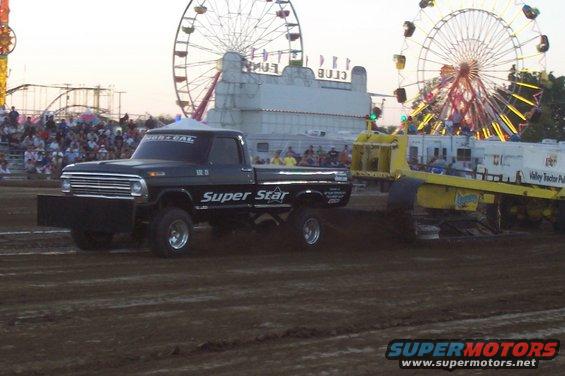 Black Ford F-Series truck with 'Super Star' on the side, pulling a sled at a fair. Ferris wheels in background