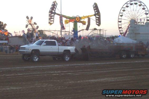 White 1994 Ford F-150 pickup truck with lifted suspension and chrome wheels pulls a sled on a dirt track at a fair