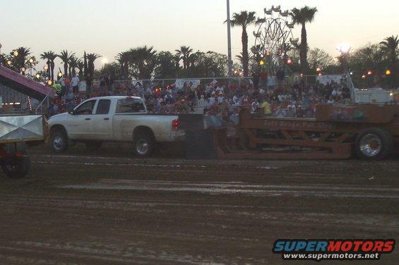 White pickup truck pulling a large trailer in a dirt arena with a crowd watching and a Ferris wheel in the background
