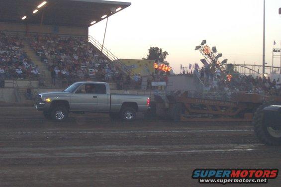 Silver Dodge Ram pickup truck pulling a sled down a dirt track with a crowd in the bleachers