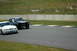 1993 Ford Mustang - Two Ford Mustangs, one modern and one classic, are on a paved race track with grassy banks and a concrete barrier.