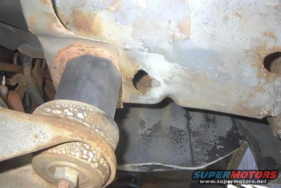 Rusted frame section with a black rubber bushing and suspension arm. A hole is visible in the frame
