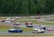 1965 Ford Mustang - Several vintage cars are racing on a track, with a wooded area visible behind the safety fence.