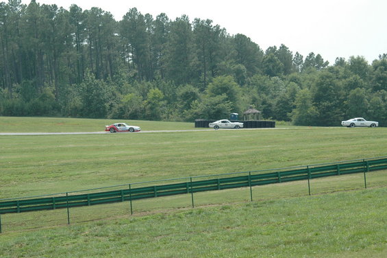 Three cars racing on a track with trees and a fence in the foreground