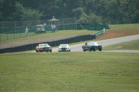 Three classic muscle cars racing on a track: orange, white with blue stripes, and black