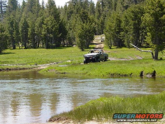 Pickup truck driving through shallow water on a dirt path, surrounded by trees and grass