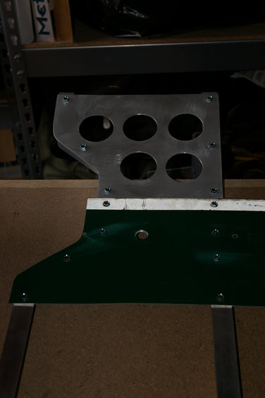 metal bracket with six circular holes, attached to a green panel with screws