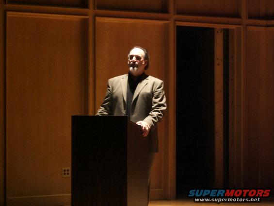 Man in a suit stands behind a podium in a room with wood-paneled walls